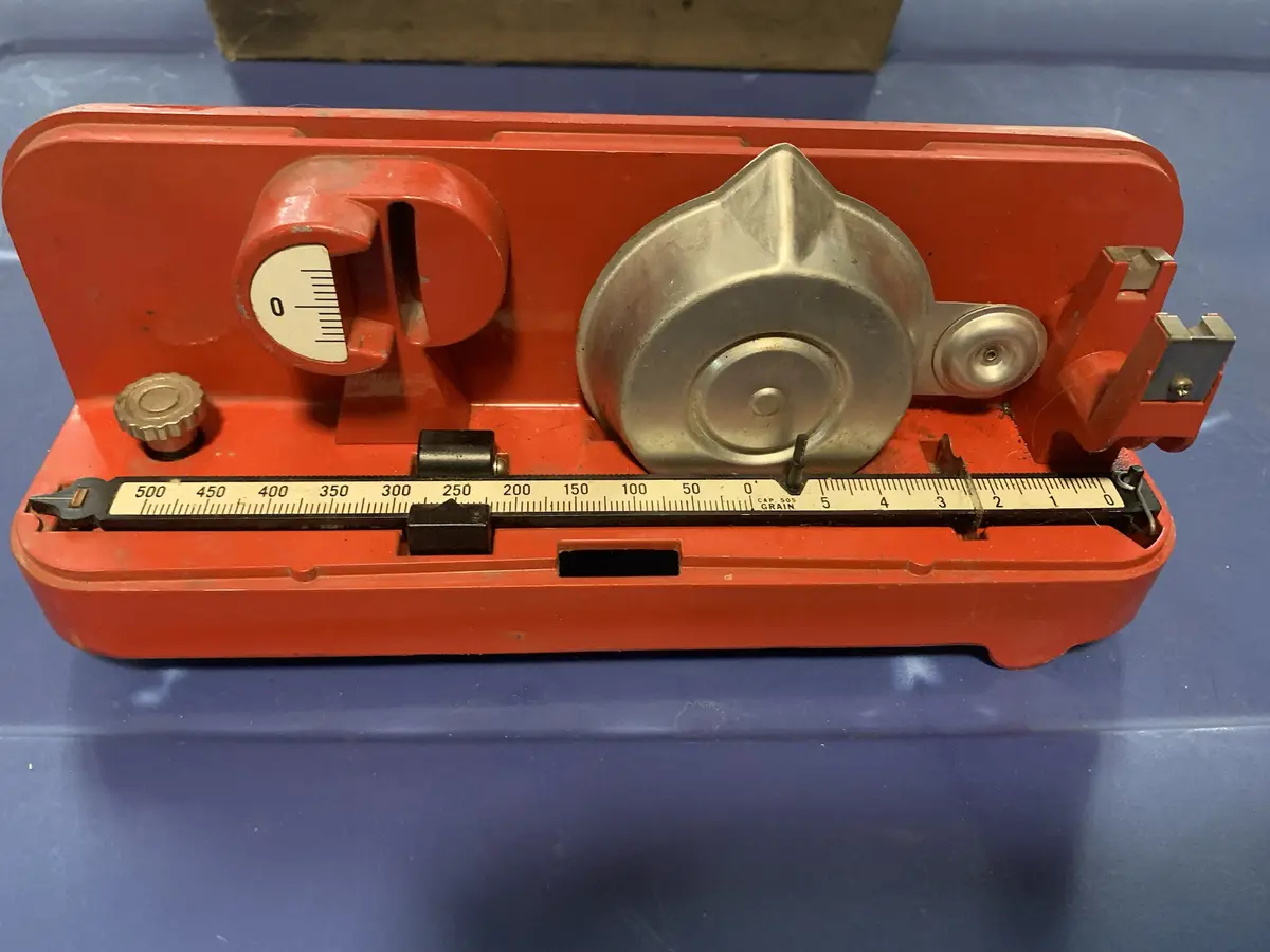 A red, vintage-style mechanical scale with a silver bowl and a sliding ruler marked in grains is shown on a blue surface…