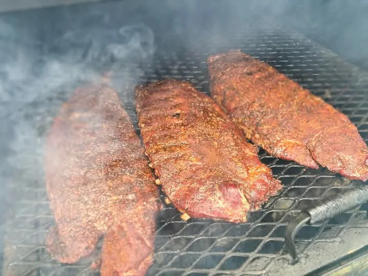 Three large, seasoned meat ribs are cooking on a smoky grill grate, with visible smoke rising around them. The overlaid text…
