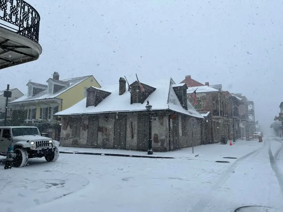 A snowy street scene in New Orleans, featuring historic buildings with snow-covered roofs and a white jeep parked on the…