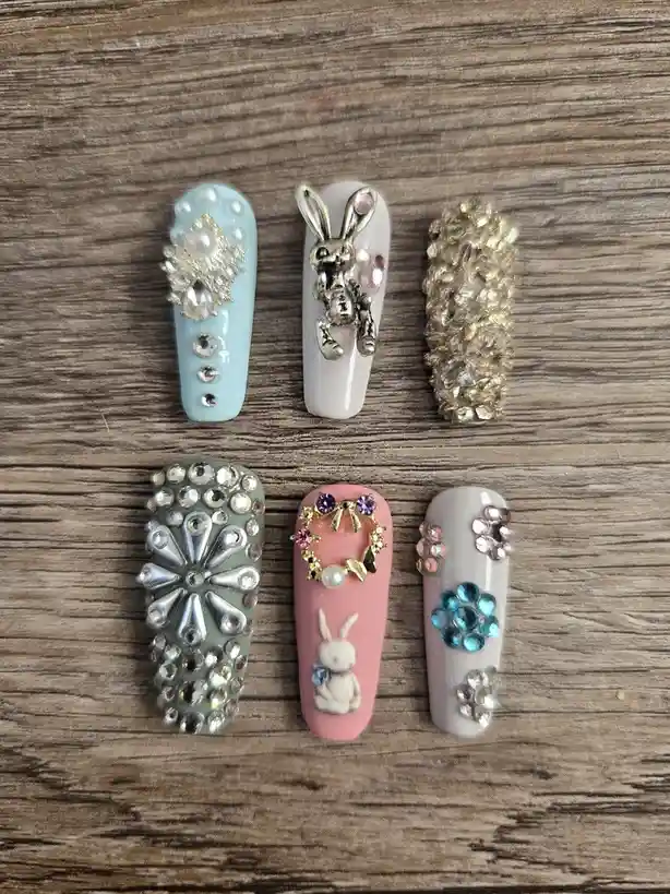 Six ornate, long acrylic fingernails with elaborate decorations are arranged in two rows on a wooden surface. Designs…