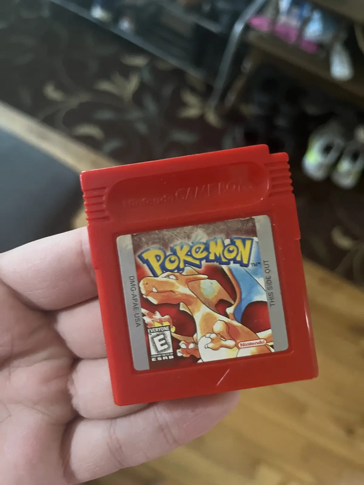 A hand holds a red Nintendo Game Boy cartridge for the Pokémon game, featuring a worn label with Charizard artwork and the…