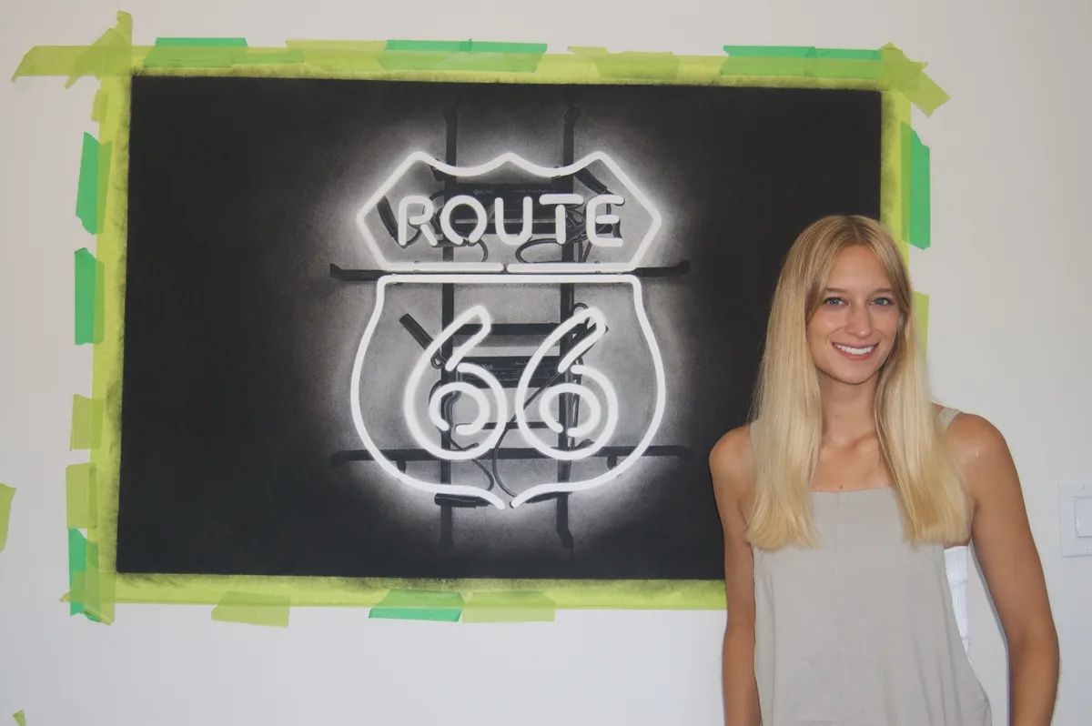 A woman with blonde hair smiles beside a black wall framed with green painter's tape, which displays a glowing white neon…