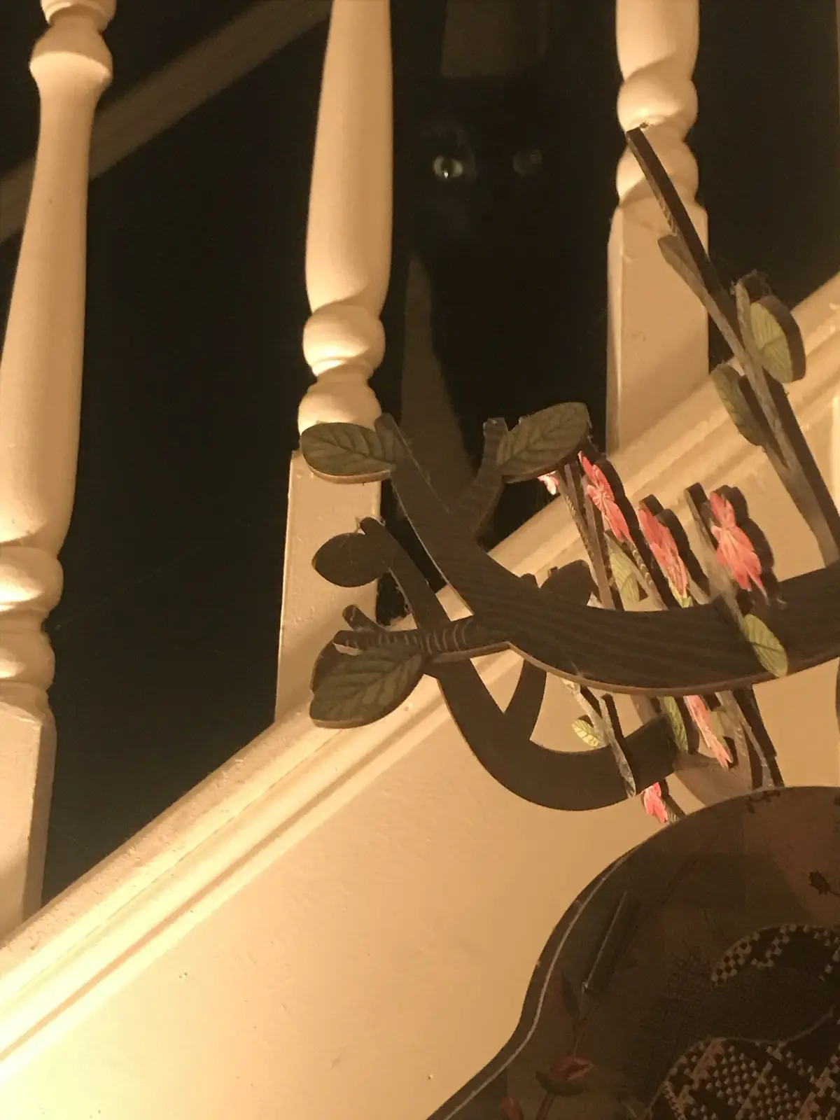A black cat's glowing eyes peer out from behind a dark staircase banister, partially obscured by decorative metalwork with…