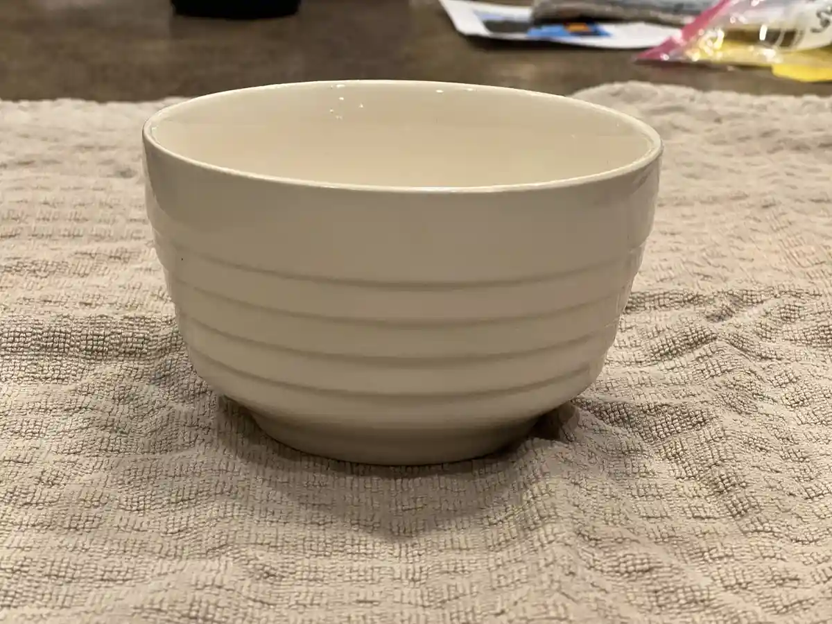 A plain white ceramic bowl with horizontal ridges sits centered on a textured beige towel on a countertop. The overlaid text…
