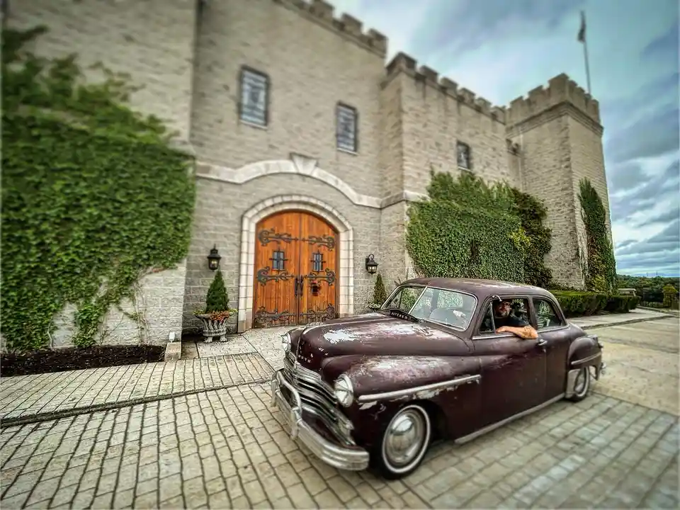 A man with a beard and glasses leans out of the driver's side window of a rusty, vintage maroon car parked on a stone…