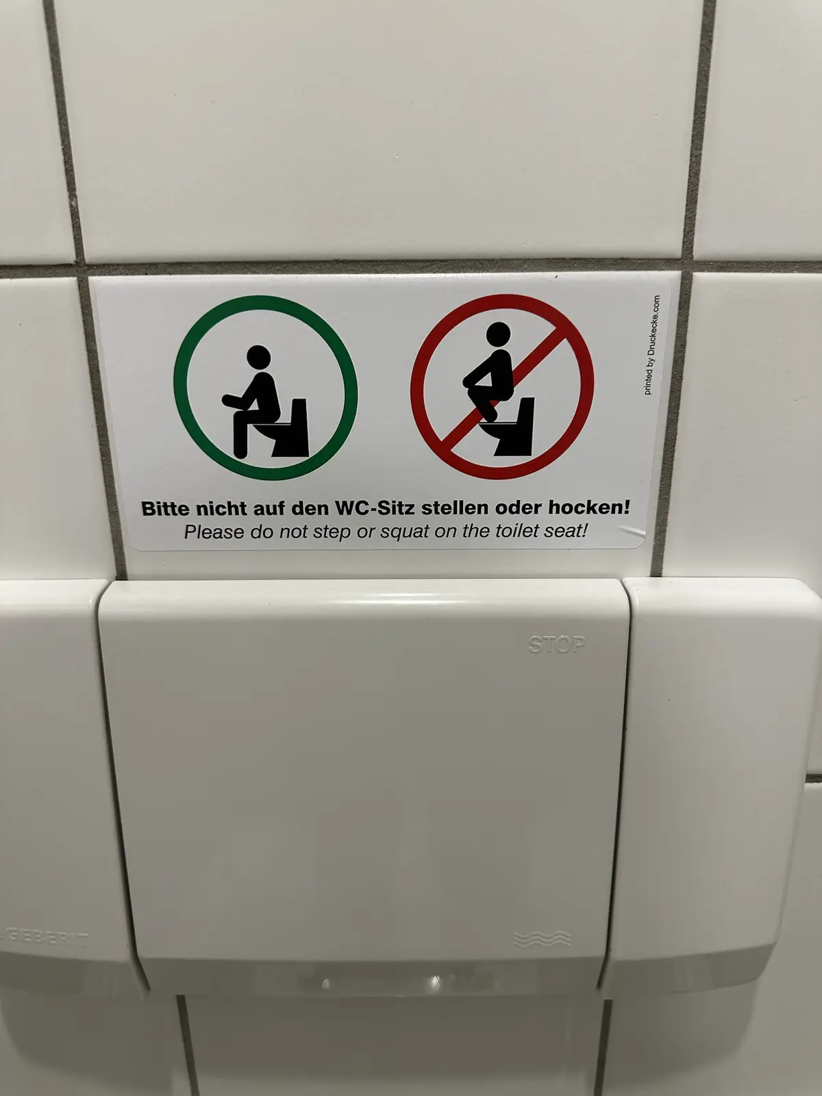 A close-up of a public restroom sign on a tiled wall, featuring two pictograms: one green circle showing a person sitting on…