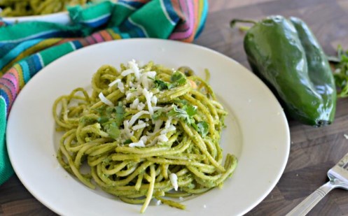 a white plate holds spaghetti coated in green pesto sauce, topped with shredded white cheese and chopped green herbs. A…