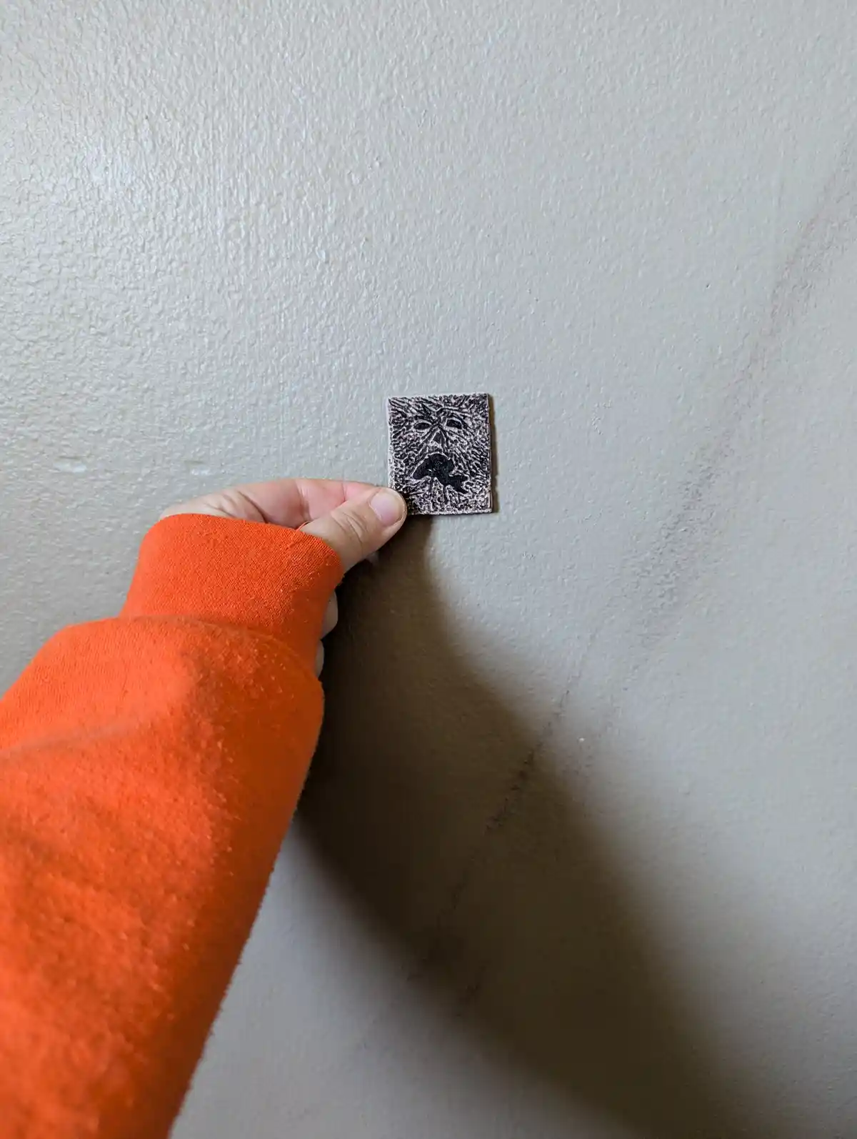 A hand in an orange sleeve holds a small square patch against a textured beige wall, the patch featuring a crudely etched or…