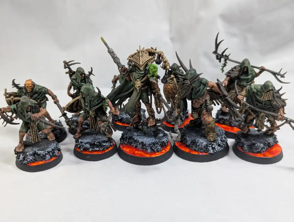 A row of eight intricately painted fantasy miniatures, depicting grotesque, green-robed warriors with horns, skulls, and…
