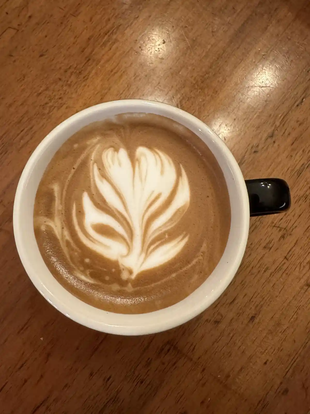 A top-down photo of a white ceramic coffee cup with a black handle, sitting on a wooden table. The coffee has intricate…