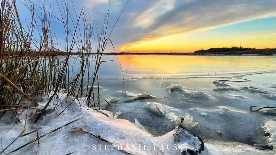A serene winter sunset over a partially frozen lake, with ice chunks and snow in the foreground and tall reeds on the left…