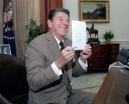 A smiling man in a suit sits in an office, holding up a piece of paper with a crude drawing of a face. He holds a pen in his…