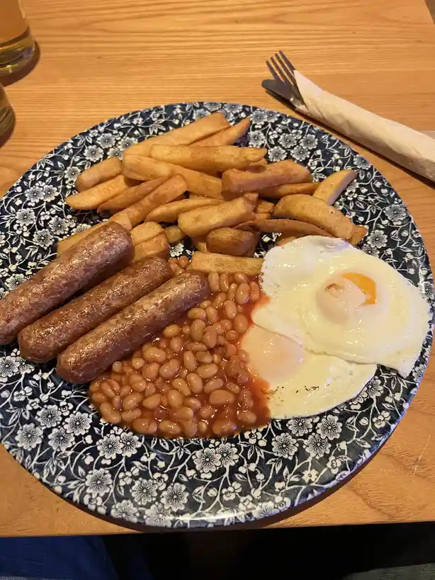A plate of traditional British breakfast food including fried eggs, baked beans, three sausages, and chips, served on a blue…