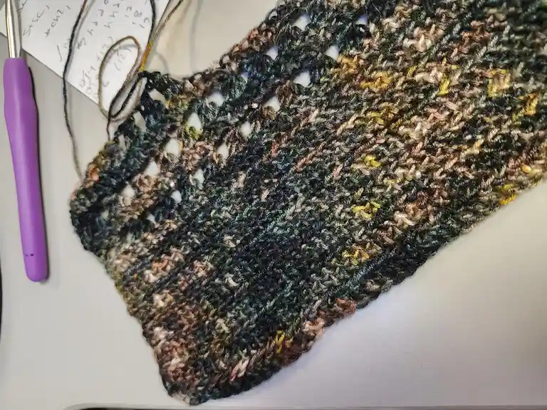 A close-up photo shows a partially completed, multicolored crocheted blanket with a messy, uneven edge, next to a purple…