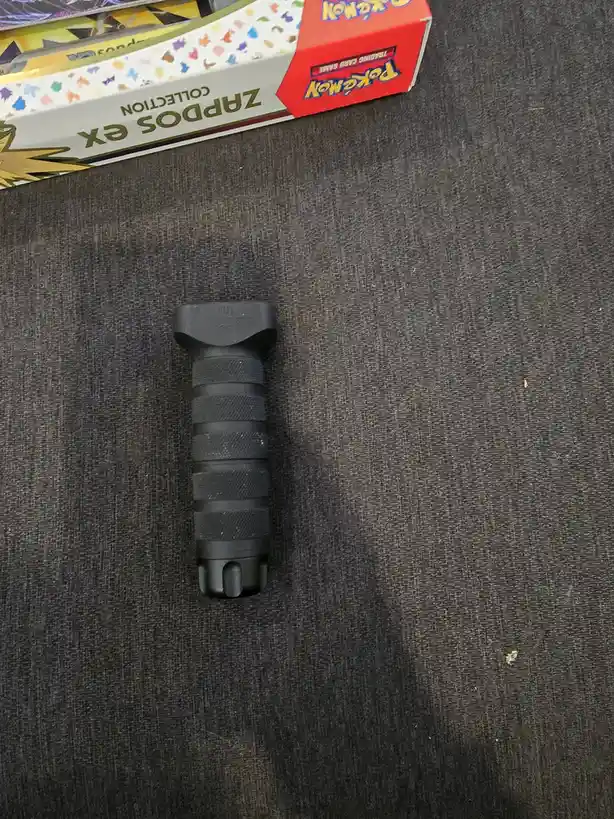 A black, textured, cylindrical object resembling a gun grip lies on a dark gray fabric surface. In the upper left corner, a…