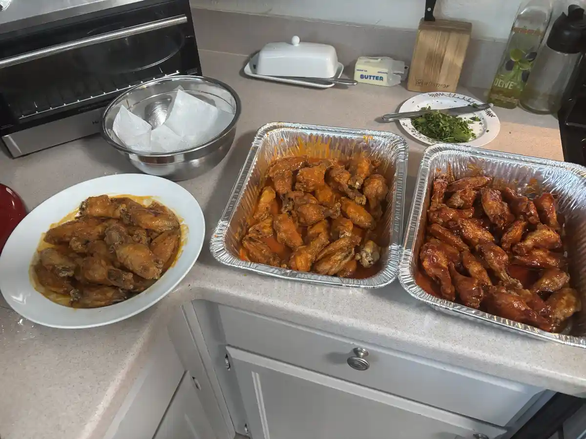 A kitchen counter displays three large aluminum trays and one white plate, all filled with saucy chicken wings. In the…