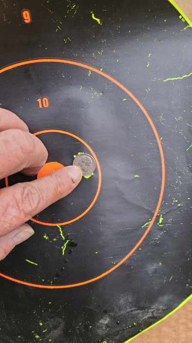 A close-up photo shows a person's hand holding a small silver coin on a black target with orange concentric circles. The…