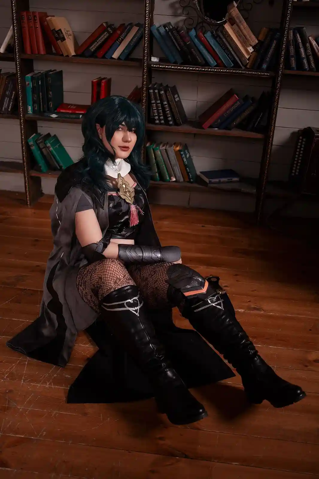 A person in a detailed cosplay of a dark fantasy character sits on a wooden floor in front of bookshelves, wearing a black…