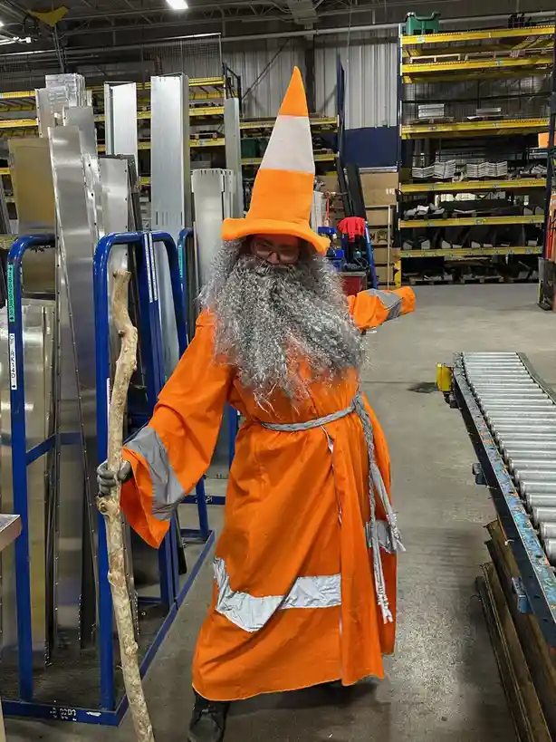A person in a warehouse wearing an orange and white traffic cone hat, an orange high-visibility robe with reflective…