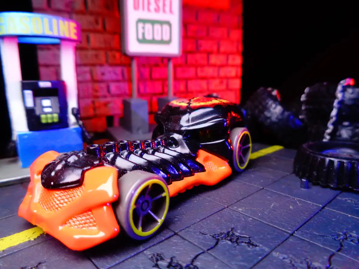 A toy car with a black and orange body and purple wheels is parked on a cracked pavement in front of a miniature gas…