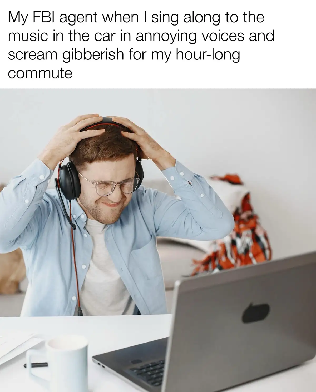 A man wearing glasses and a light blue button-down shirt sits at a desk with a laptop and a mug, holding his head with both…