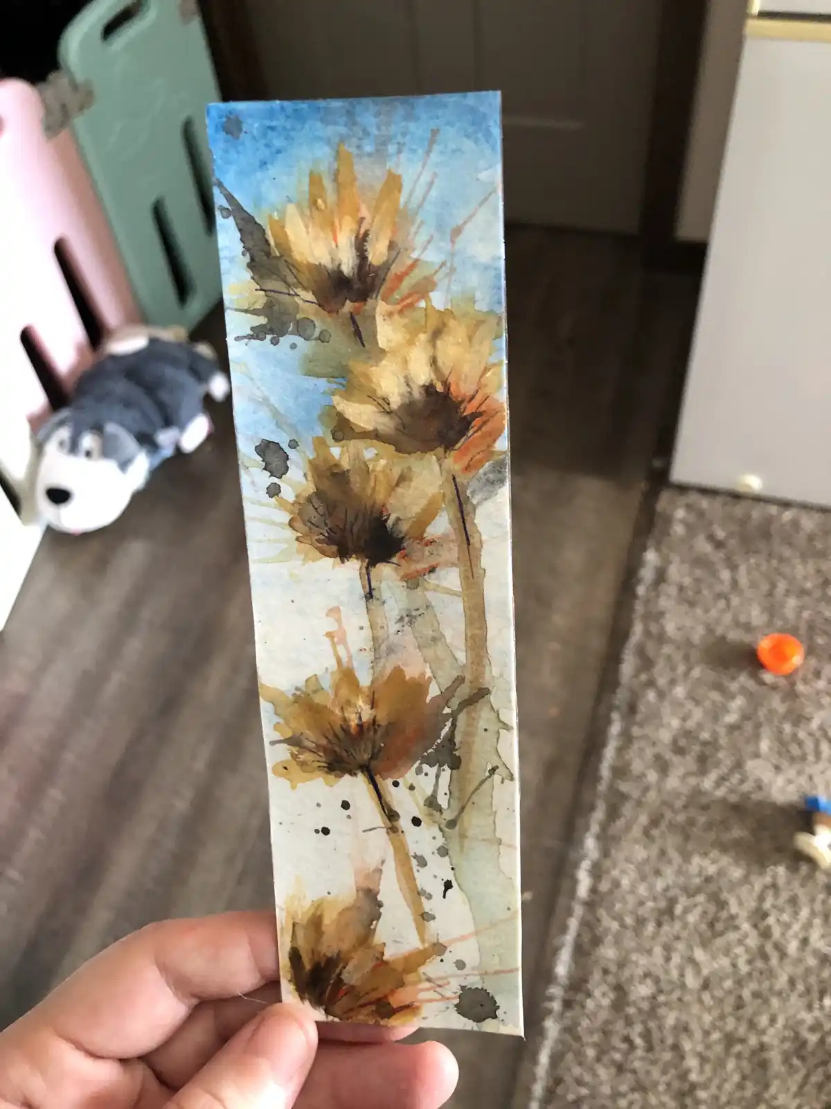 A hand holds up a small, rectangular watercolor painting depicting several stylized, brownish-yellow flowers against a blue…