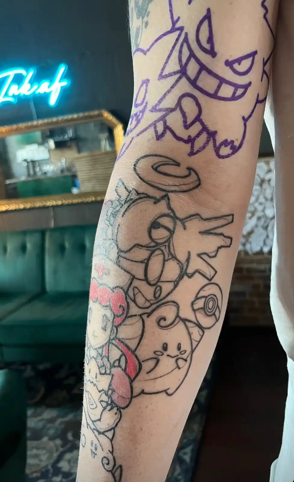 A close-up of a person's forearm showing a partially completed tattoo with purple outlines of Gengar at the top, followed by…