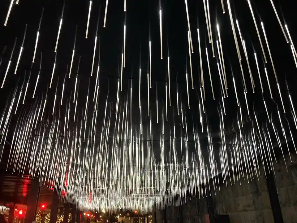 A nighttime photo shows numerous vertical, glowing white light tubes suspended from above, resembling falling rain against a…