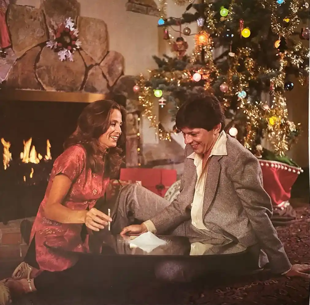 A man and woman sit on the floor in front of a fireplace and a decorated christmas tree, smiling at each other while holding…