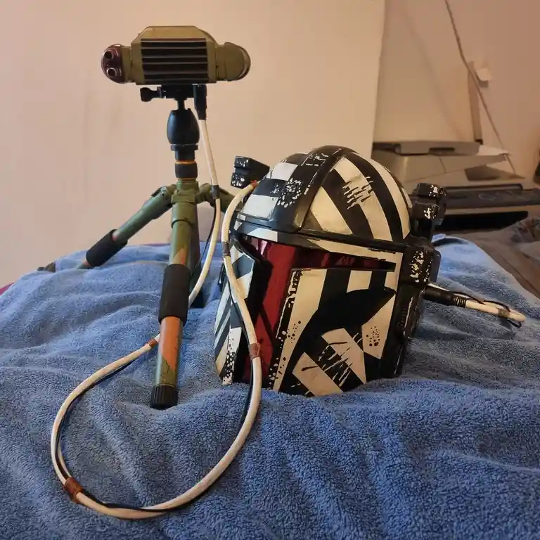 A custom-painted Mandalorian helmet with black and white geometric patterns and a red visor sits on a blue towel, connected…