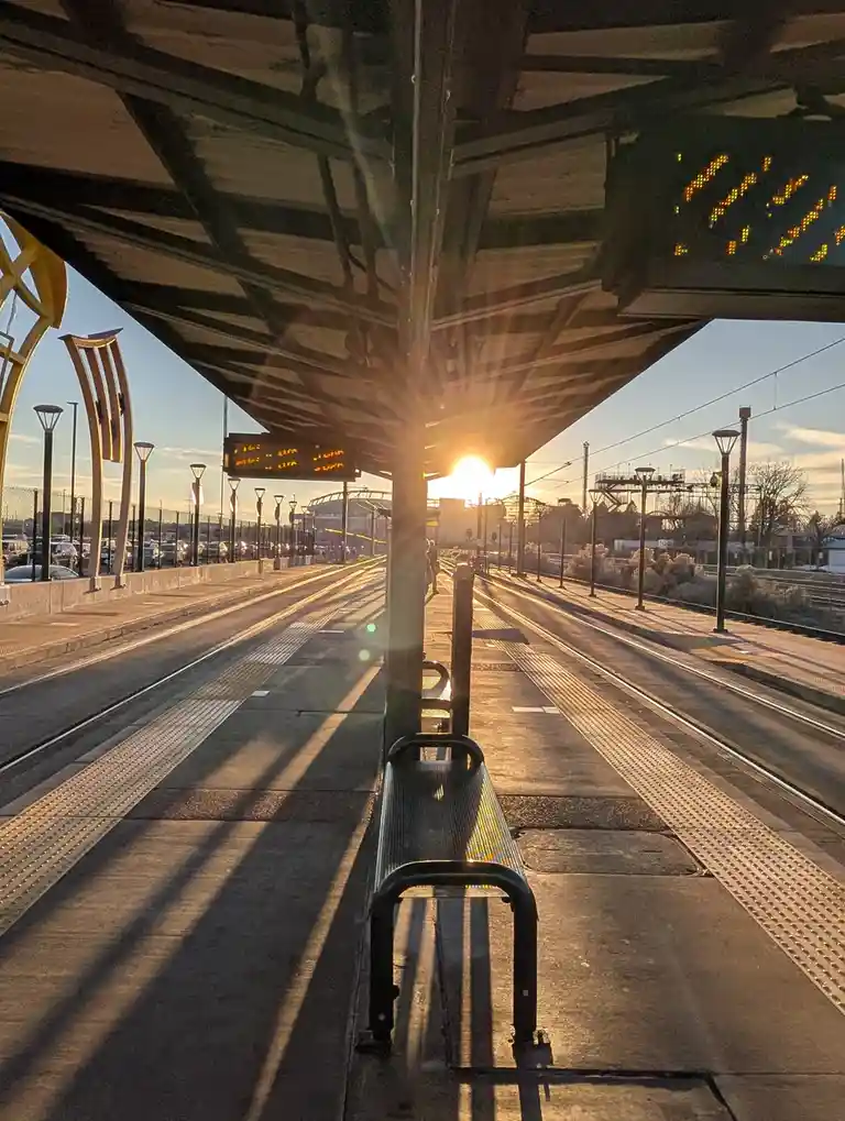 A photo of a train station platform at sunset, with the sun low in the sky casting long shadows. A metal bench sits in the…
