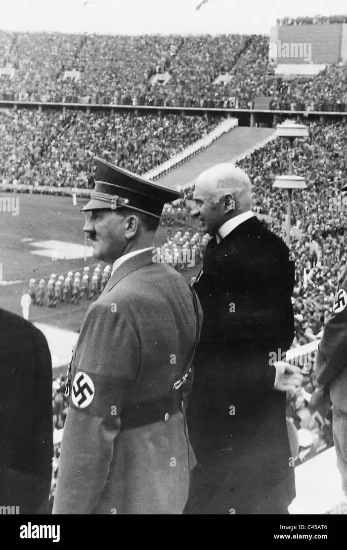 A black-and-white historical photograph shows Adolf Hitler in a military uniform with a swastika armband standing beside a…