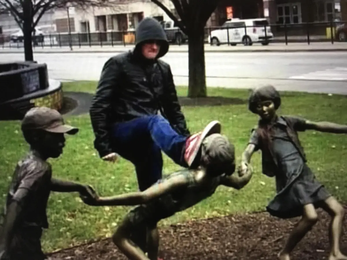 A person in a dark hoodie and jacket stands in a park next to bronze statues of children playing, with one leg raised as if…