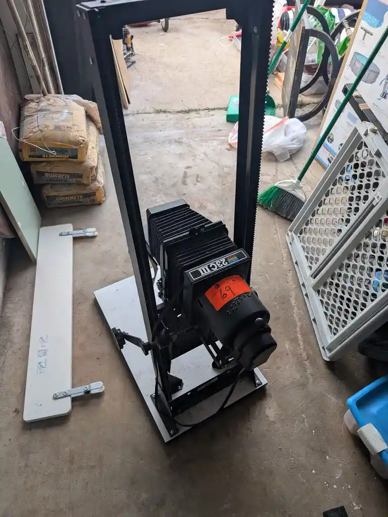 A black, vintage-style large-format camera, labeled '23C III', sits on a metal stand in a cluttered garage. An orange price…