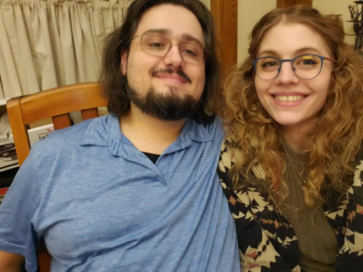 A man with long dark hair, glasses, and a beard wearing a blue polo shirt sits in a wooden chair, smiling slightly while…