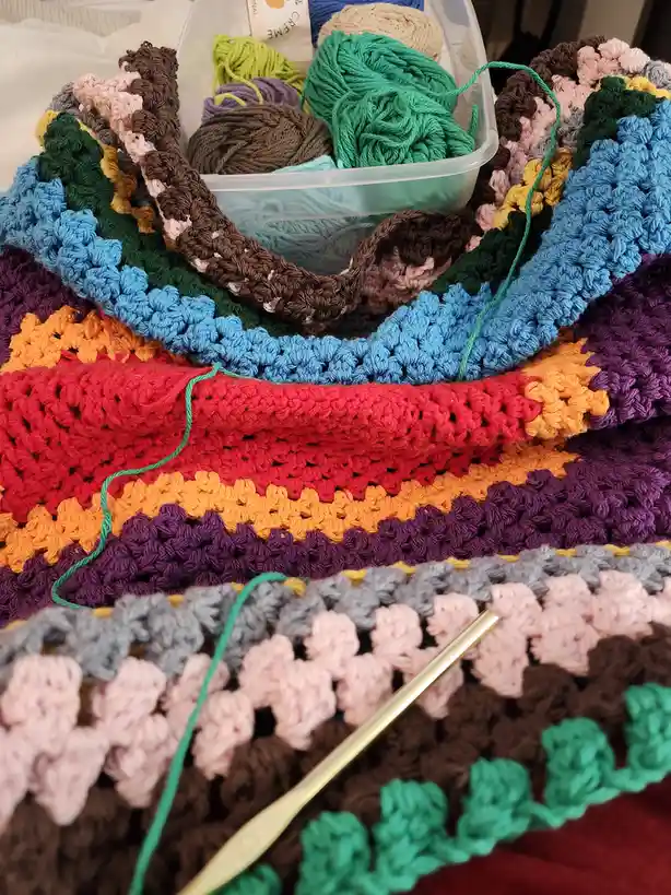 A colorful, multi-layered crochet blanket made of vibrant stripes in red, orange, purple, blue, green, and brown is shown…