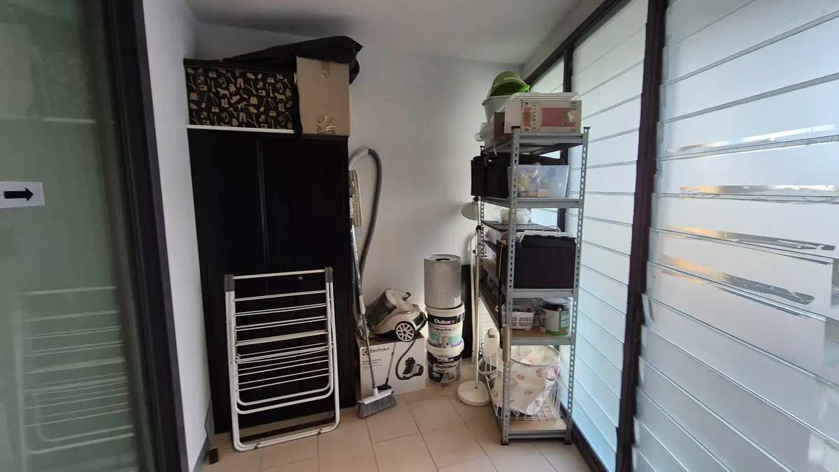 A narrow hallway or utility space filled with cluttered storage, including a black wardrobe, a metal shelving unit stacked…