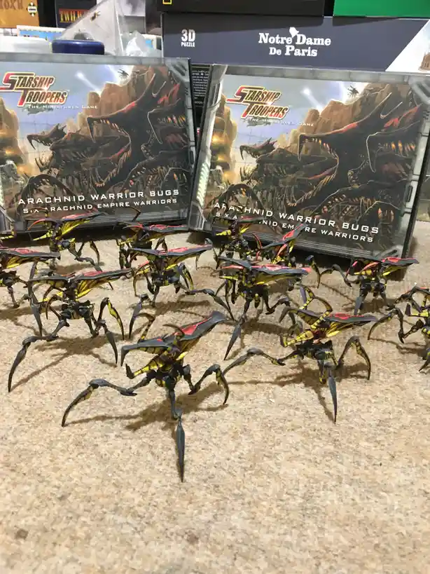 A collection of painted Starship Troopers Arachnid Warrior Bug miniatures are arranged on a textured surface, with their…