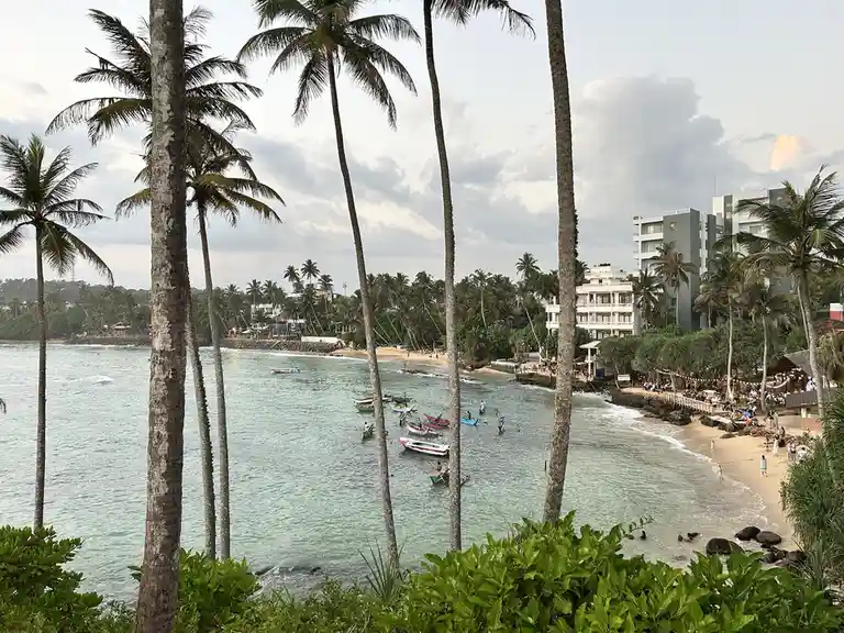 A scenic tropical beach view framed by tall palm trees, showing turquoise water, small boats, a sandy shore with people, and…