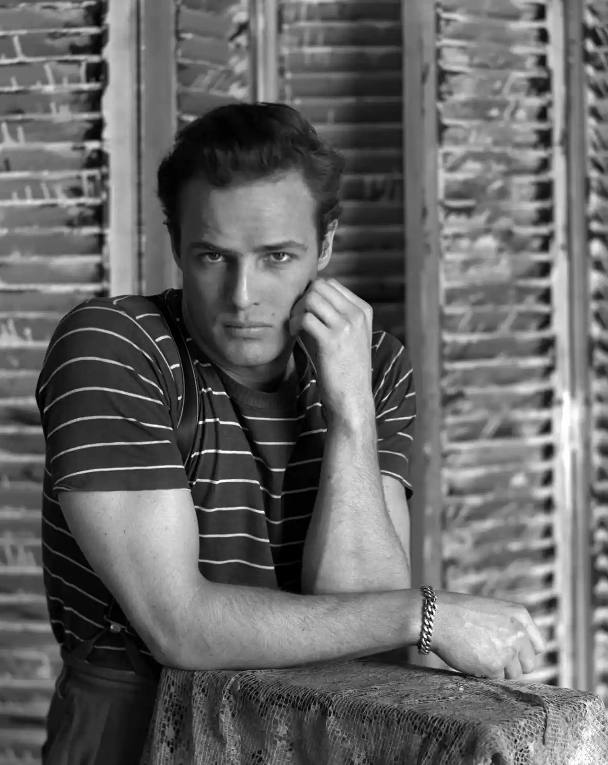 A black-and-white photo of a man in a striped shirt leaning on a table with a serious, intense gaze, his hand resting on his…