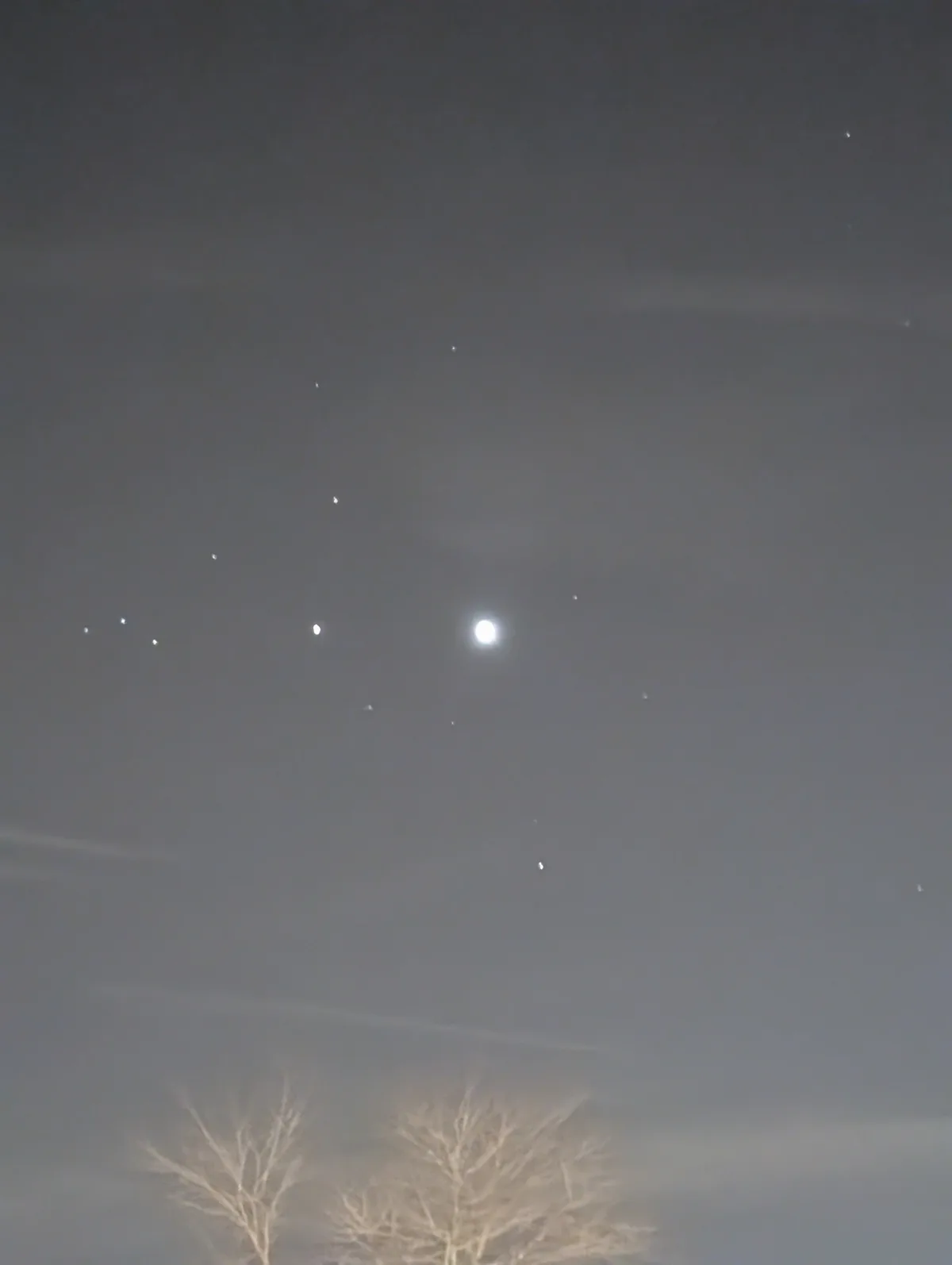 A night sky photo showing a bright moon surrounded by faint stars, with bare tree branches visible at the bottom. The…