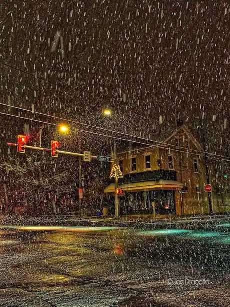 A nighttime street scene under heavy snowfall, with red traffic lights glowing, street signs including 'Linden St' and 'ONE…