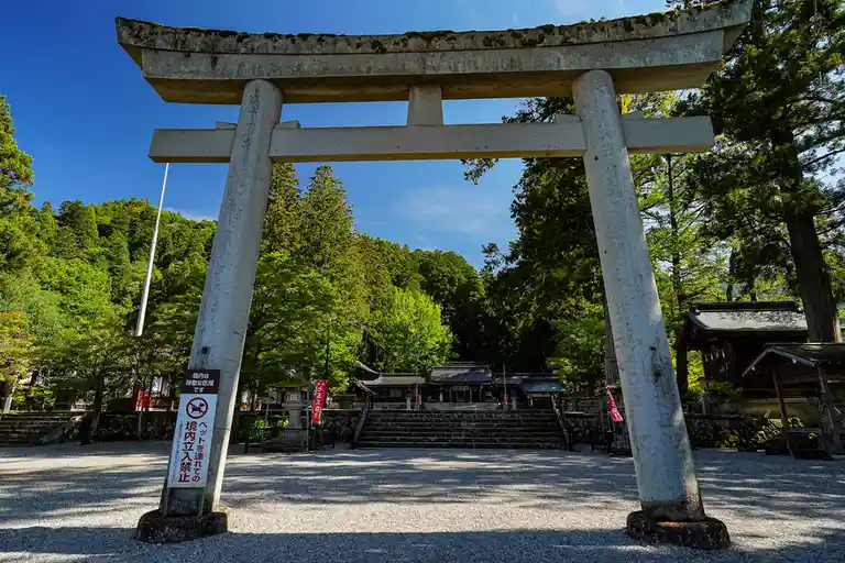 A large, weathered stone torii gate stands at the entrance to a Japanese shrine, framed by lush green trees and a clear blue…