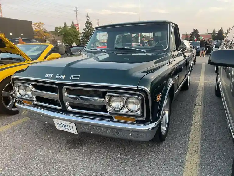 A dark green classic GMC pickup truck is parked in a lot, its Ontario license plate reads 'I'M IN IT'. The truck has a…
