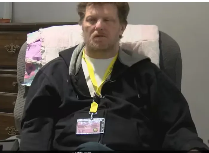 A man with a beard and messy hair sits in a chair wearing a black jacket over a white shirt and a yellow lanyard with an ID…