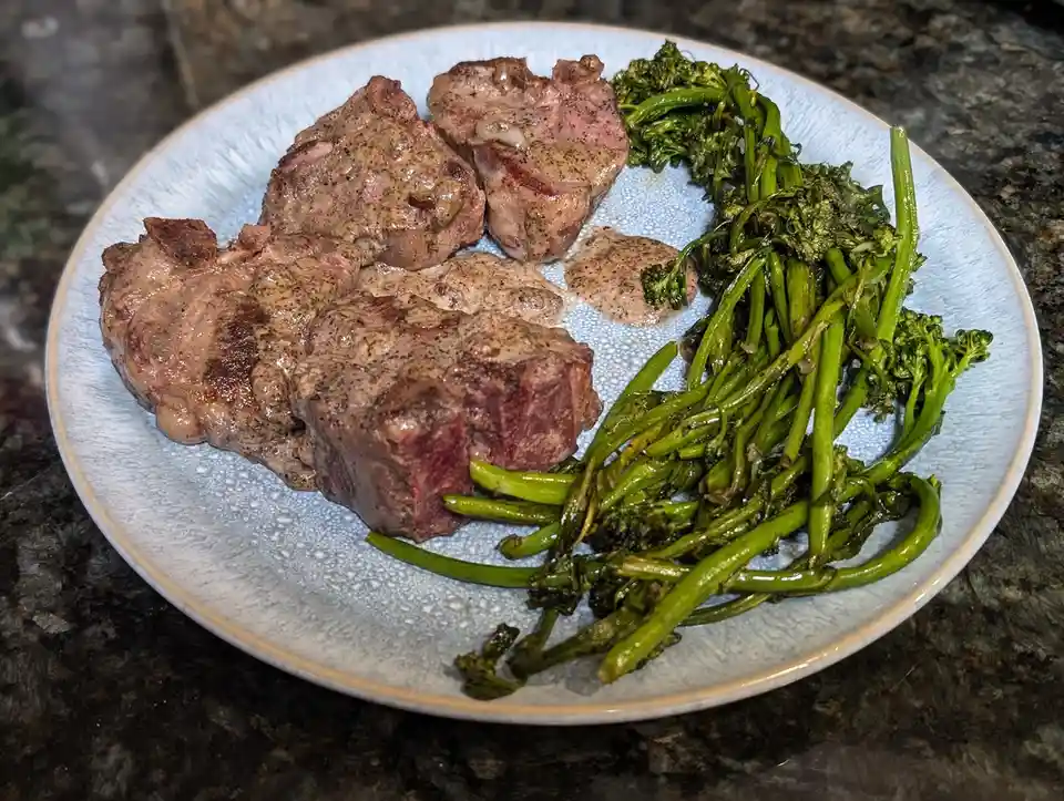 A plate of cooked steak with a brown sauce and roasted broccolini on a light blue textured plate, set on a dark granite…