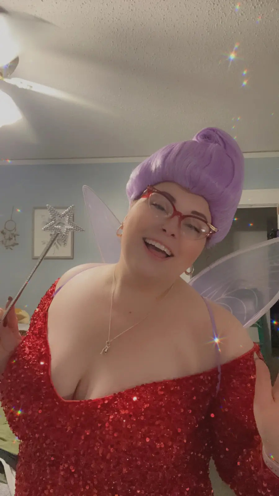 A person with purple hair in a bun, wearing red-rimmed glasses and a sparkly red off-the-shoulder top, poses with…