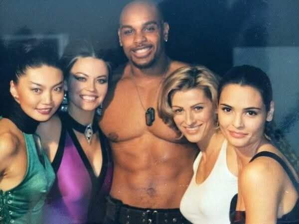 A shirtless man with a goatee and necklace stands center, flanked by four smiling women in evening wear. The overlaid text…