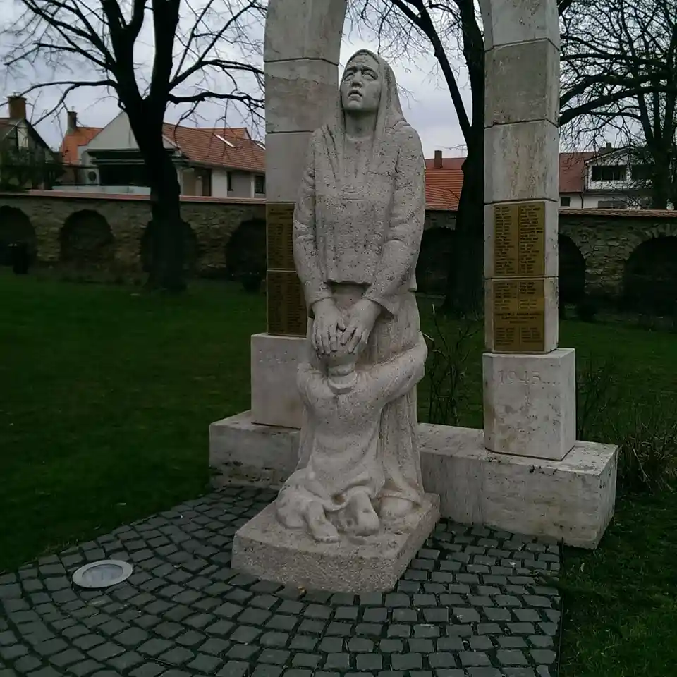 A somber stone statue depicts a woman in a headscarf holding a child, standing between two pillars marked '1941-' and…