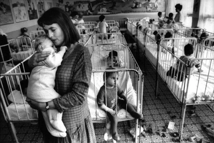 A black-and-white photo shows a woman holding a baby in a nursery filled with rows of cribs, some occupied by children. The…